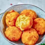 A bowl containing six golden-brown fried macaroni and cheese balls, one of which is bitten open to reveal the creamy filling inside.
