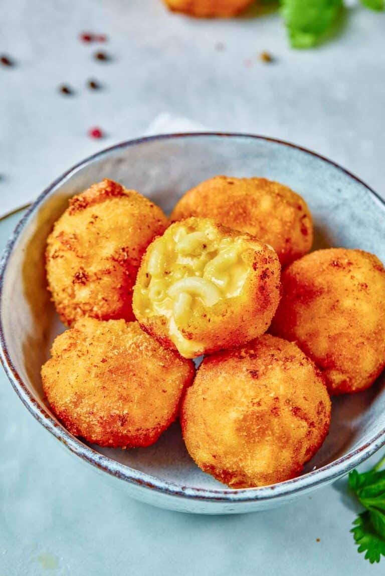 A bowl containing six golden-brown fried macaroni and cheese balls, one of which is bitten open to reveal the creamy filling inside.
