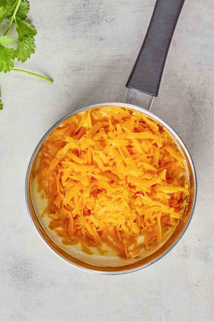 A saucepan containing a creamy white sauce topped with shredded cheddar cheese sits on a light countertop. Some green herbs are visible in the upper left corner.