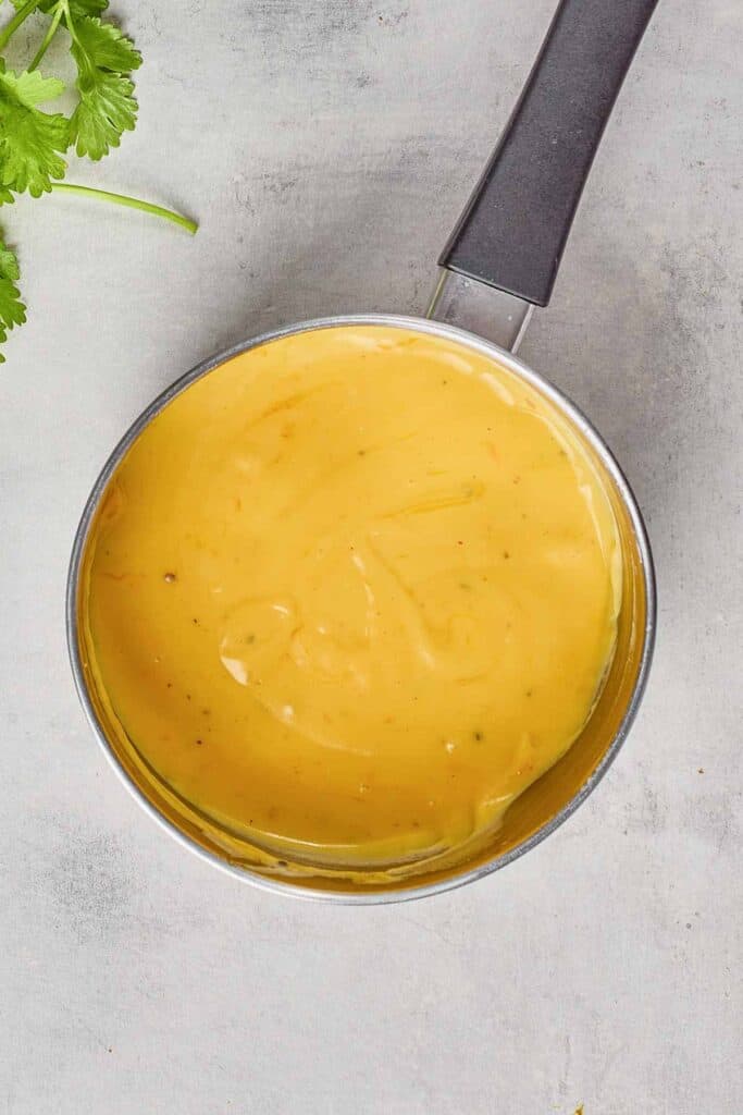 A small saucepan filled with creamy, yellow cheese sauce sits on a light gray surface. Some green herb leaves are visible in the top left corner.