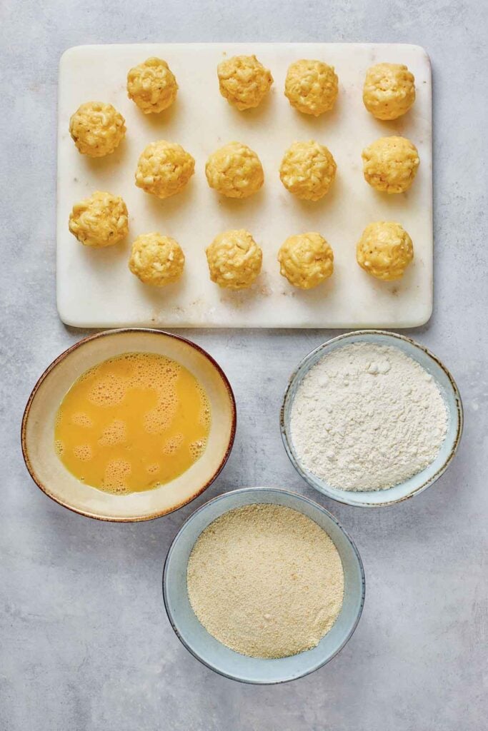 Fourteen uncooked cheese balls on a white board, with bowls of beaten eggs, flour, and breadcrumbs arranged below on a light surface.