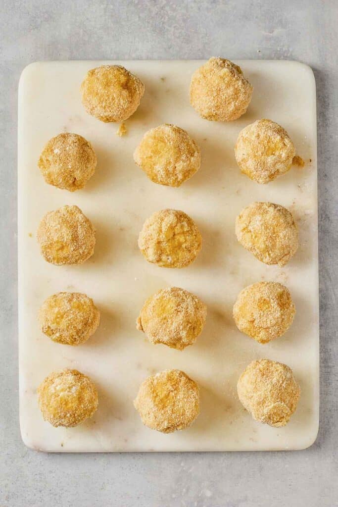 Fifteen round, breaded balls are evenly spaced in a grid pattern on a white rectangular cutting board, viewed from above.