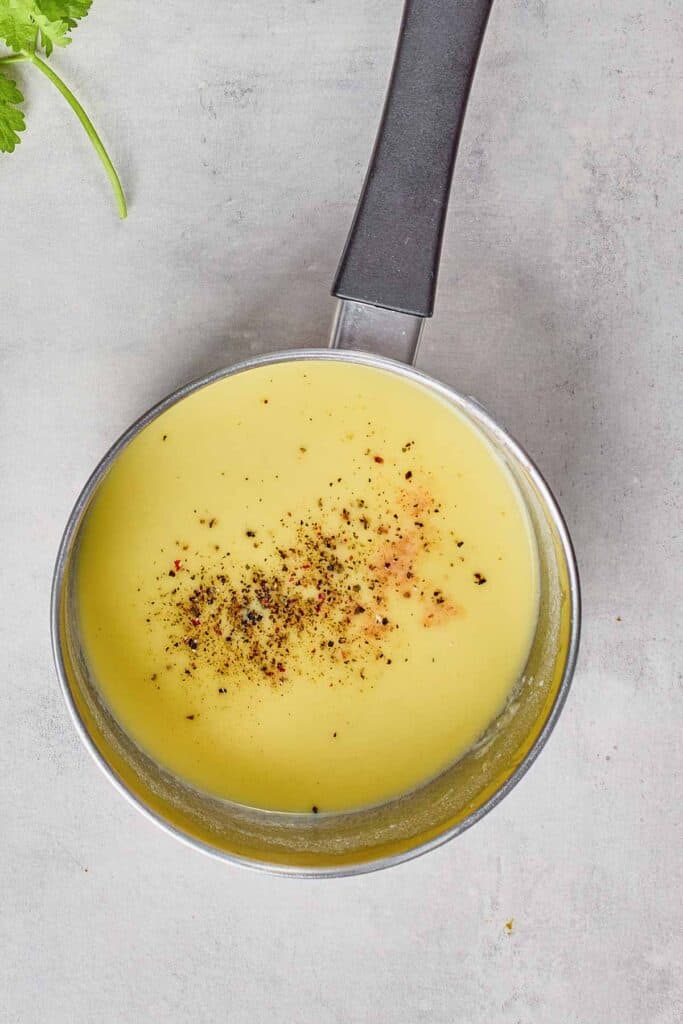 Small saucepan filled with creamy yellow sauce, topped with cracked black pepper, placed on a light gray surface.