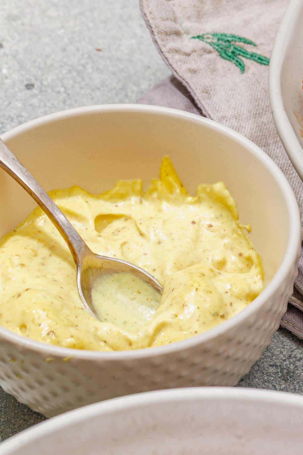 A white bowl filled with creamy yellow sauce or dip, with a metal spoon resting inside.