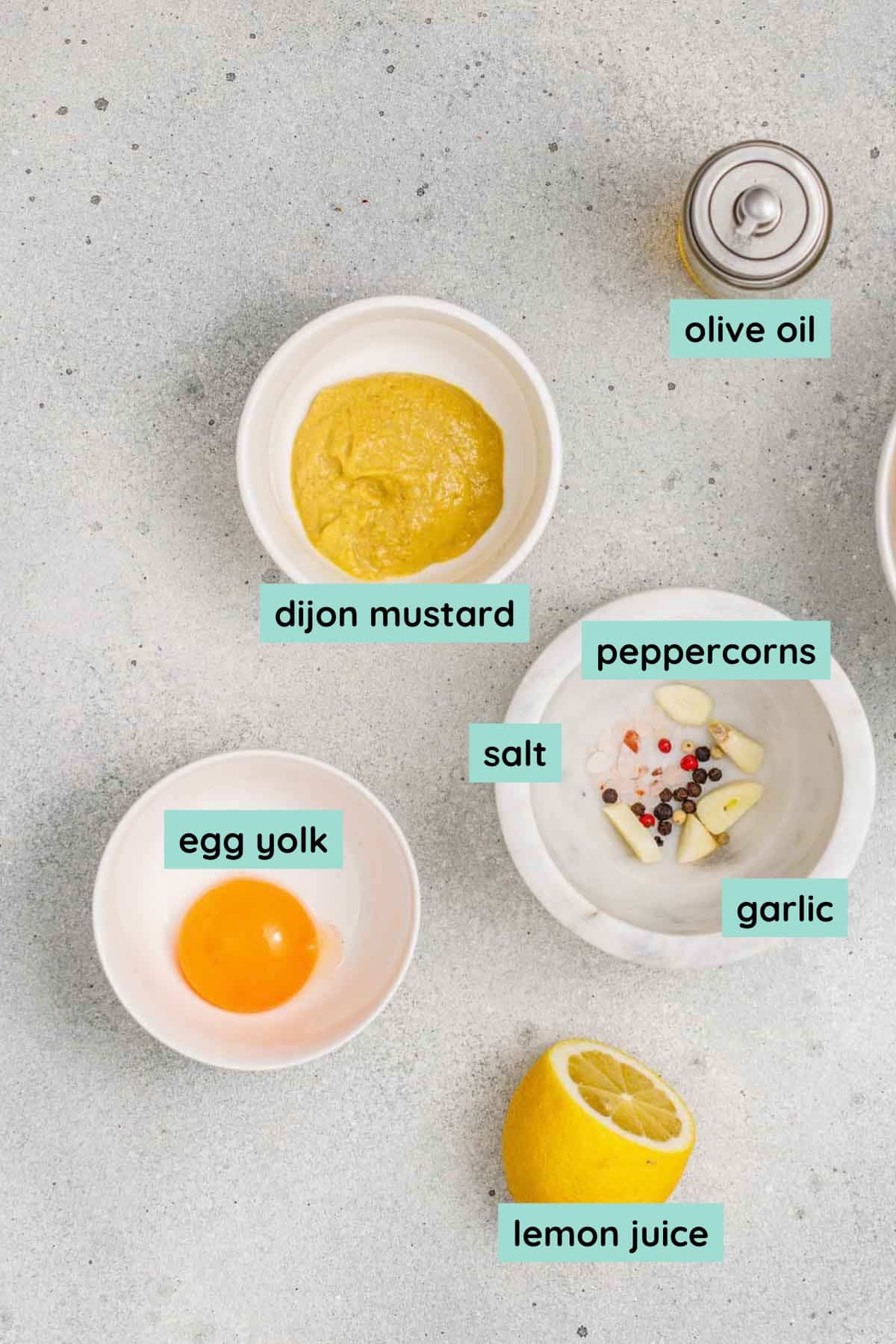 Bowls containing dijon mustard, egg yolk, garlic, salt, peppercorns, and a bottle of olive oil, with a halved lemon and labeled ingredient names.