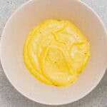 A beige bowl filled with a thick yellow sauce or dip sits on a light gray textured surface.