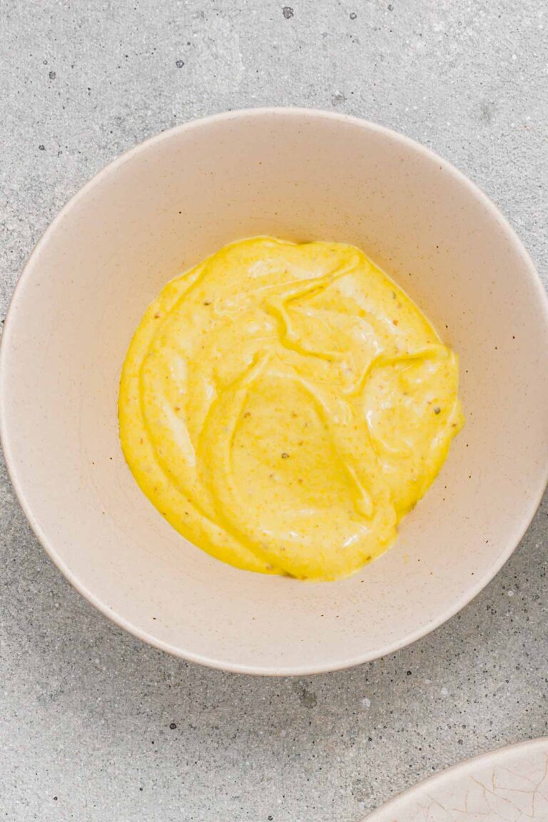 A beige bowl filled with a thick yellow sauce or dip sits on a light gray textured surface.