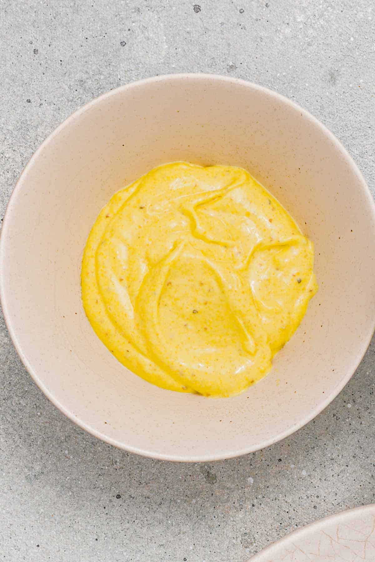 A beige bowl filled with a thick yellow sauce or dip sits on a light gray textured surface.