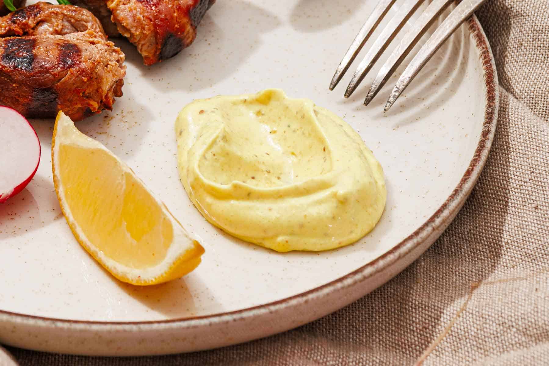 A plate with a lemon wedge, a dollop of yellow sauce, grilled meat, a radish slice, and a fork on a beige napkin.