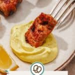 Grilled meat is dipped into a swirl of homemade aioli on a plate, with a lemon wedge and a knife nearby.