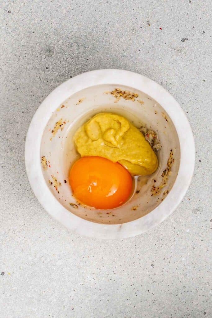A white bowl contains a raw egg yolk and a dollop of yellow mustard on a gray countertop.