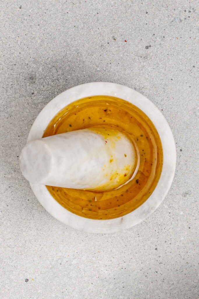 A white marble mortar and pestle containing golden yellow liquid with herbs, placed on a light gray surface.
