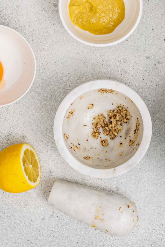 A marble mortar and pestle with crushed spices, a halved lemon, and two bowls, one with a yellow sauce, on a light countertop.