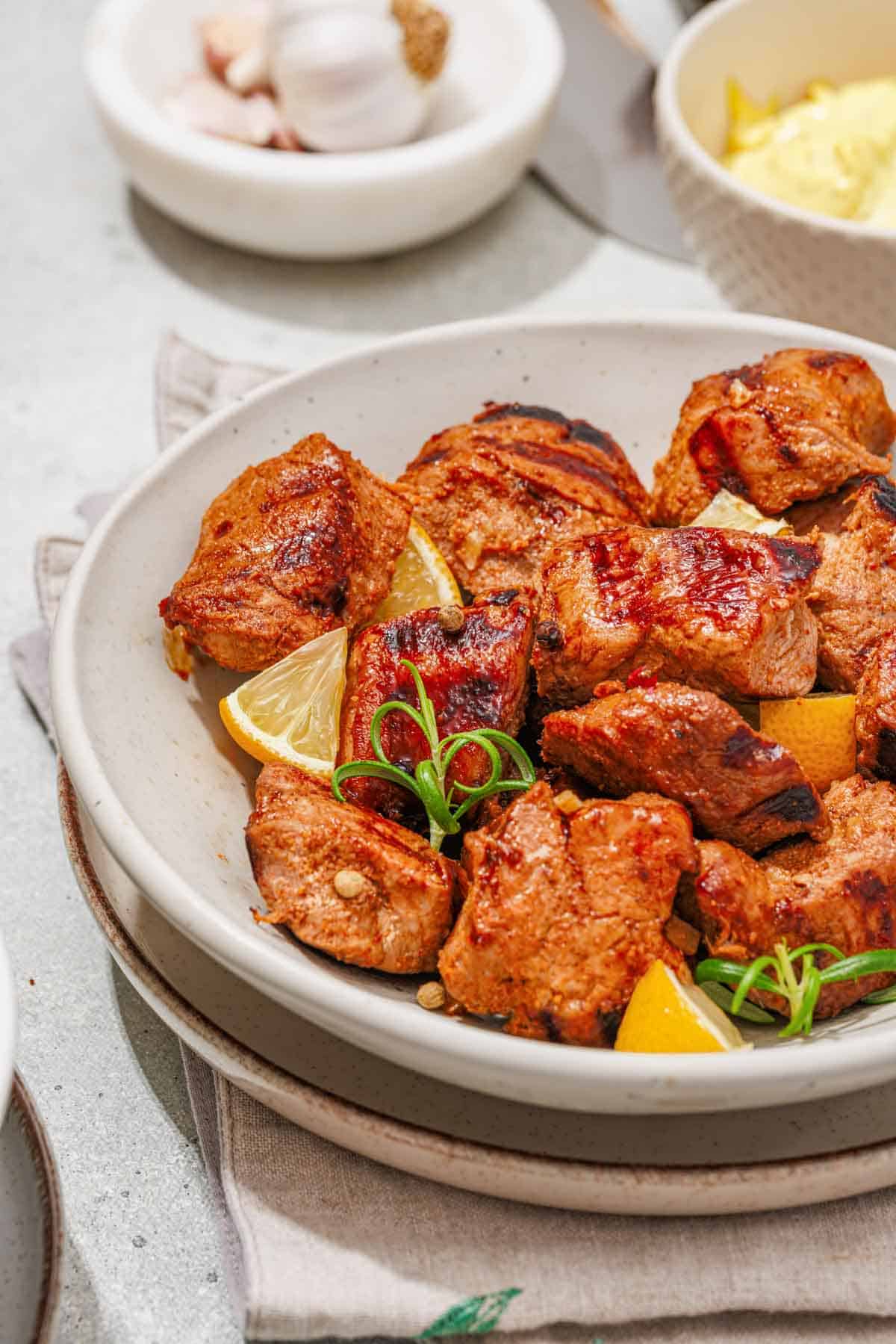A bowl of grilled meat chunks garnished with lemon wedges and herbs, with small bowls of sauce and spices in the background.