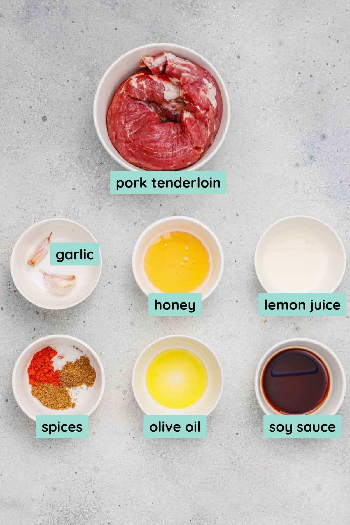 Six labeled bowls containing pork tenderloin, garlic, honey, lemon juice, assorted spices, olive oil, and soy sauce, arranged on a light gray surface.