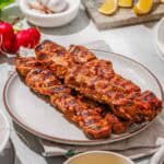 Grilled skewers of seasoned meat on a plate, surrounded by lemon wedges, garlic, radishes, and a bowl of yellow sauce on a light table.