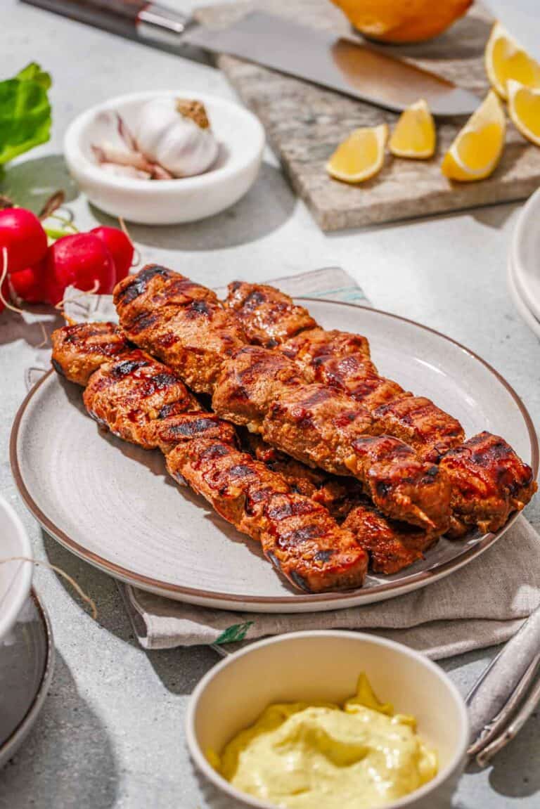 Grilled skewers of seasoned meat on a plate, surrounded by lemon wedges, garlic, radishes, and a bowl of yellow sauce on a light table.