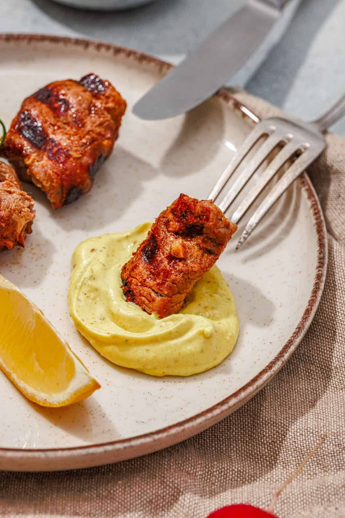 A fork holds a piece of grilled meat dipped in yellow sauce on a plate, next to a lemon wedge and another piece of meat.