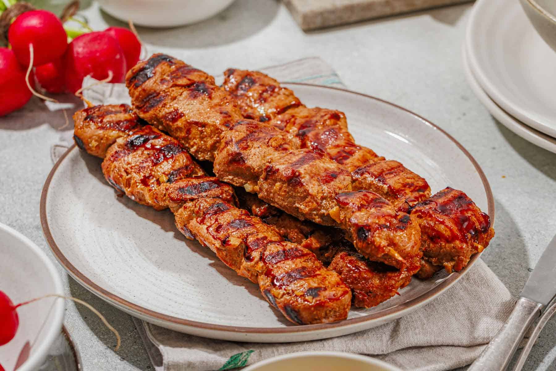 Grilled meat kebabs stacked on a white plate, with char marks visible, placed on a cloth napkin. Radishes and other dishes are partially visible nearby.