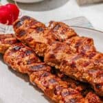 Three grilled pork skewers with a reddish marinade are arranged on a plate. Sliced lemons and a bowl of garlic are visible in the background. Text reads “Pork Skewers” at the bottom.