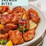 A bowl of grilled marinated pork skewer bites garnished with lemon wedges and herbs, placed on a neutral-colored plate with a website URL at the bottom.