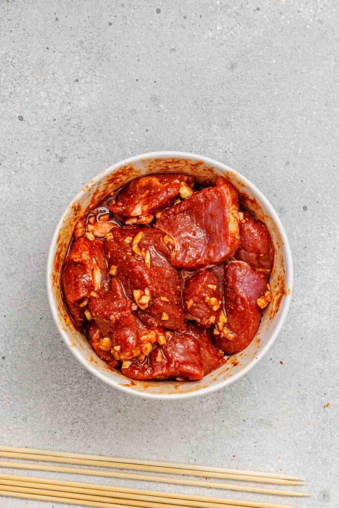 Bowl of raw marinated meat chunks with chopped garlic and spices on a gray surface, next to a few wooden skewers.