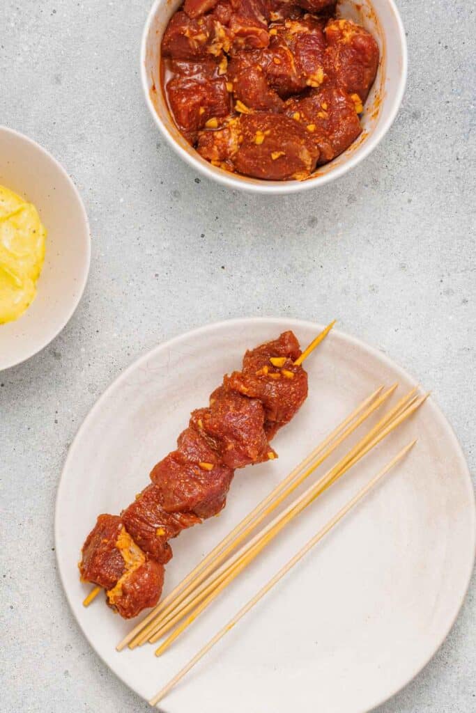 Plate with a raw marinated meat skewer and several empty wooden skewers, next to a bowl of marinated meat and a partial view of a plate with pineapple.